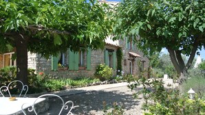 Un edificio de piedra con contraventanas verdes, un camino de grava y una mesa metálica blanca con sillas.