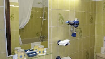 A bathroom with green tiles, a glass shelf with toiletries, a shower, and a toilet paper holder.