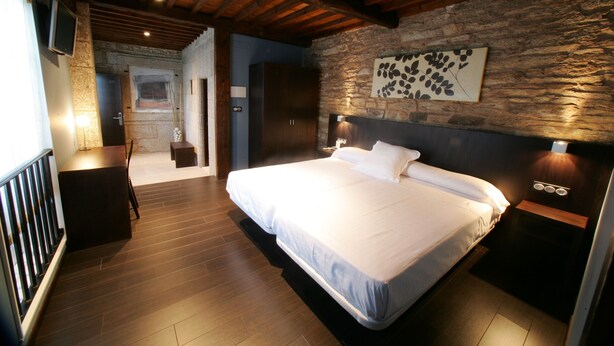 A bedroom with a large bed, wooden floor, stone wall, and a mounted TV. - A Tafona do Peregrino (Santiago de Compostela)