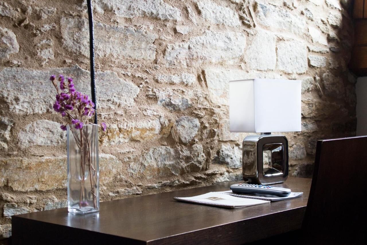 Un escritorio de madera con un jarrón transparente con flores moradas, un cuaderno, un bolígrafo y un teléfono inteligente. Una lámpara y un pequeño dispositivo electrónico están sobre el escritorio. El fondo muestra una pared de piedra texturizada.