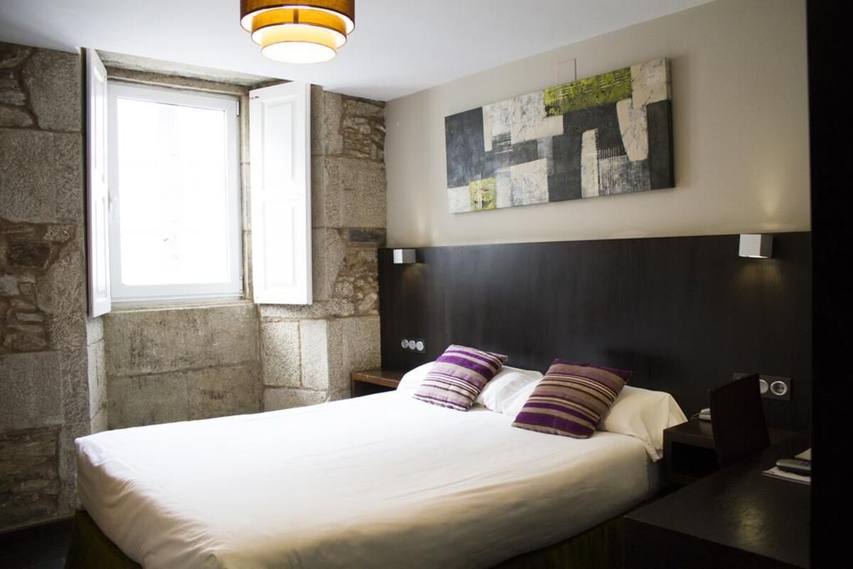 Un dormitorio con cama, ventana, pared de piedra, lámpara moderna y una obra de arte abstracto en la pared.