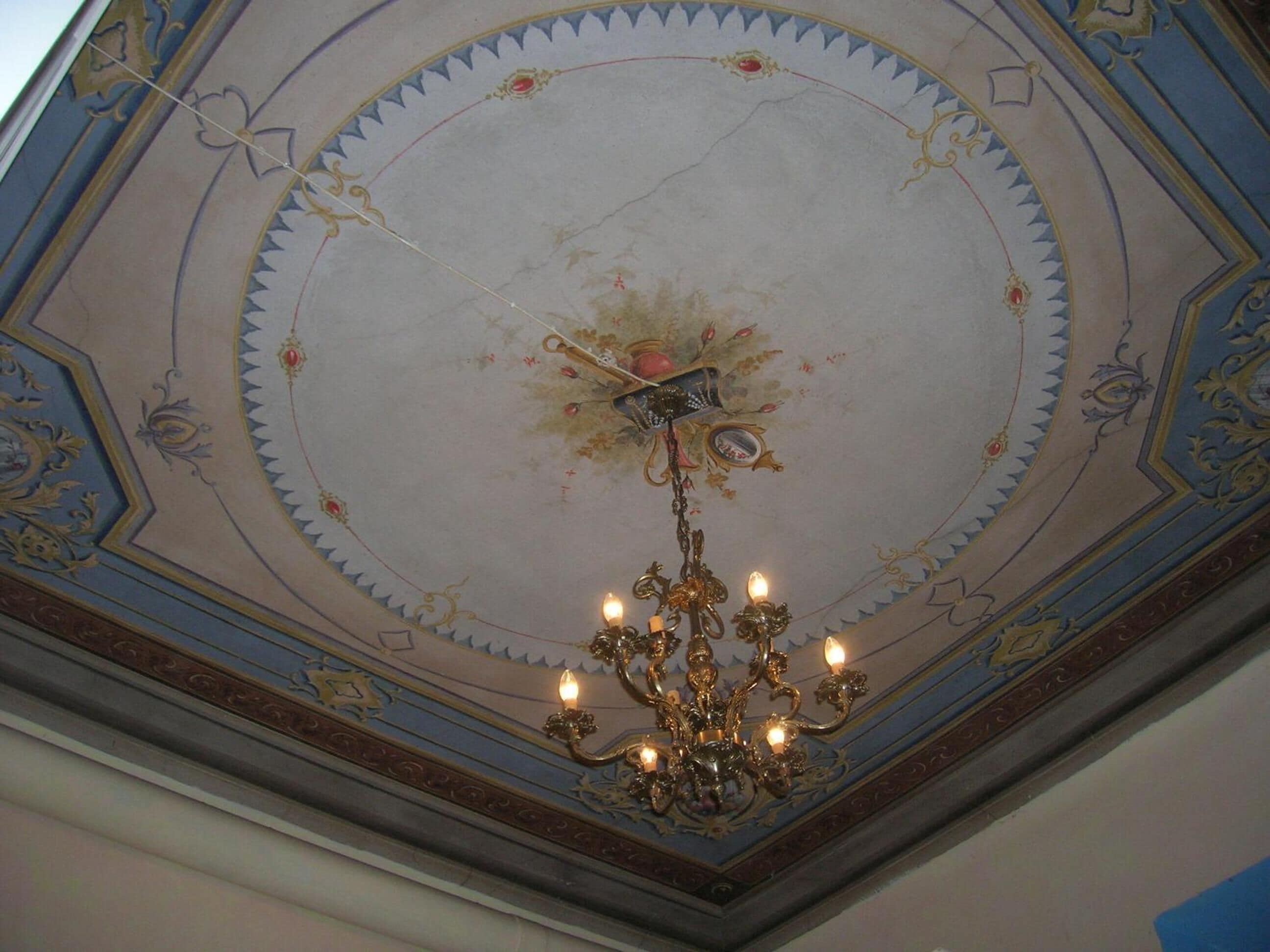 Een plafond met gedetailleerde gouden en blauwe patronen, een kroonluchter met meerdere lampen en een centraal decoratief element.