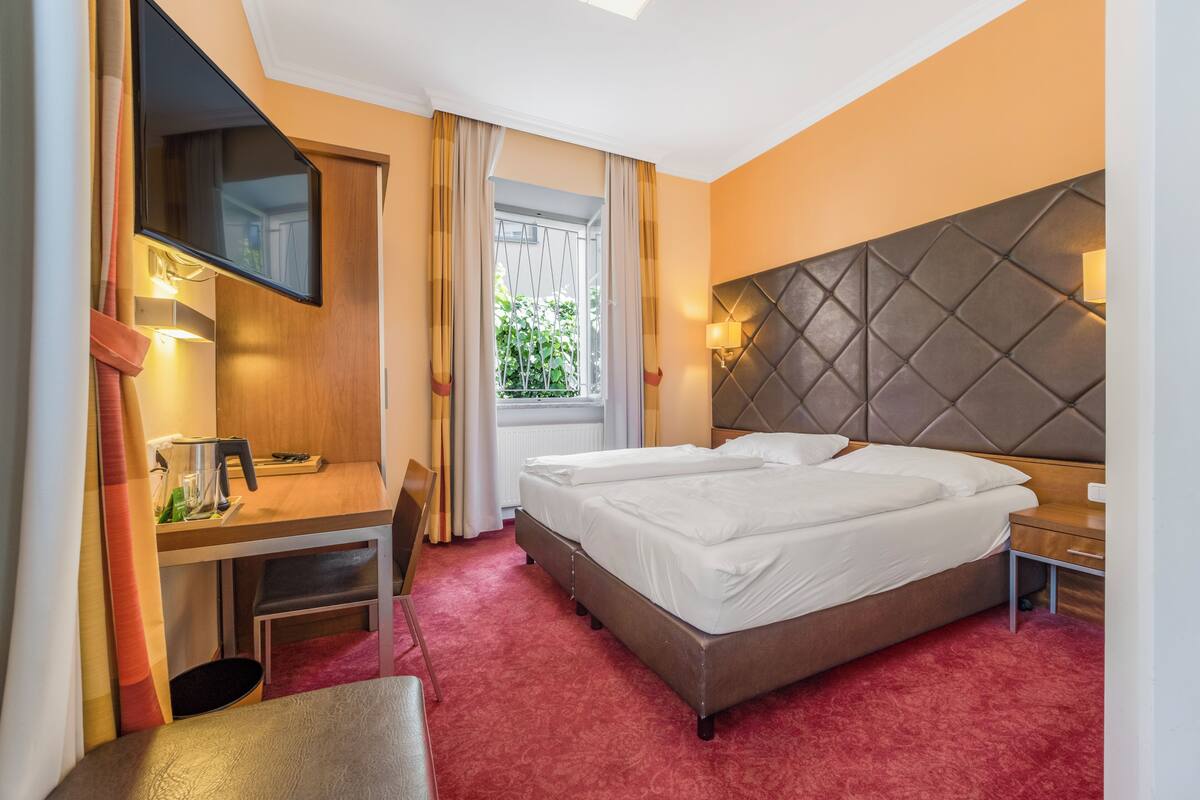Une chambre d’hôtel avec un lit, un bureau, une chaise, une télévision et une fenêtre avec des rideaux.
