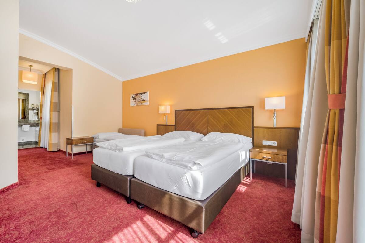 Une chambre d’hôtel avec un lit double, des tables de chevet, une commode, un miroir et une salle de bain visible à travers une porte ouverte.