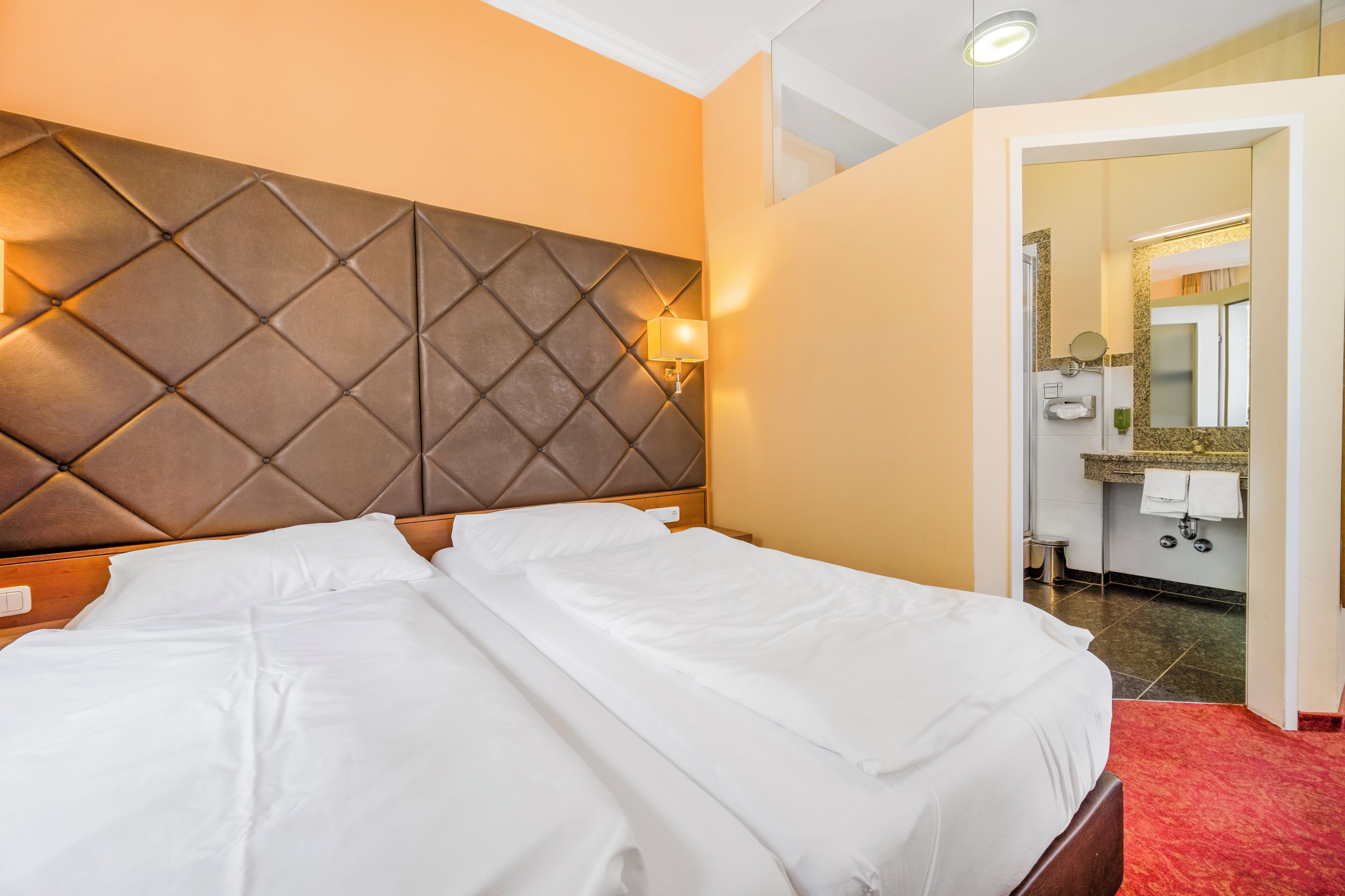 A hotel room with a large bed, a headboard with a light fixture, a bathroom visible through an open door, and a red carpet.