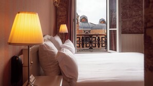 A hotel room with a bed, a nightstand, a lamp, and a view of a building through a window. - Hotel Regyn's Montmartre (Paris)