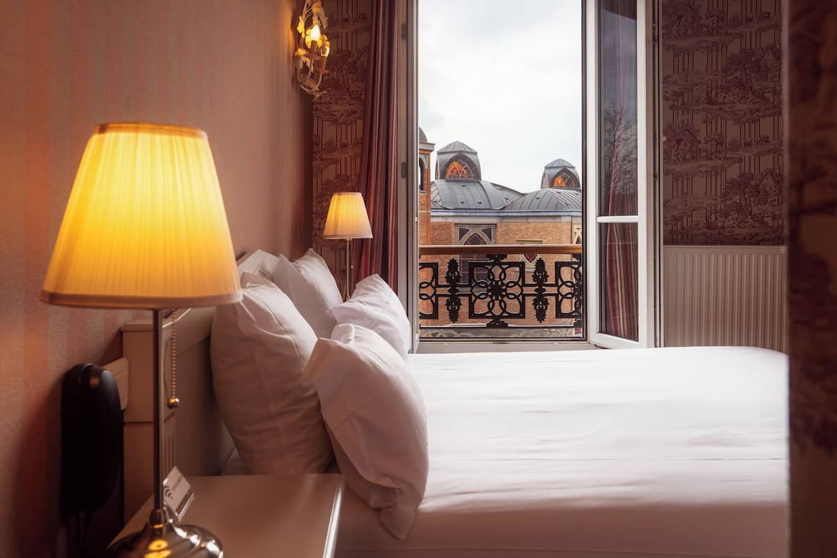 Een hotelkamer met een bed, nachtkastje, lamp en uitzicht op een gebouw door het raam.