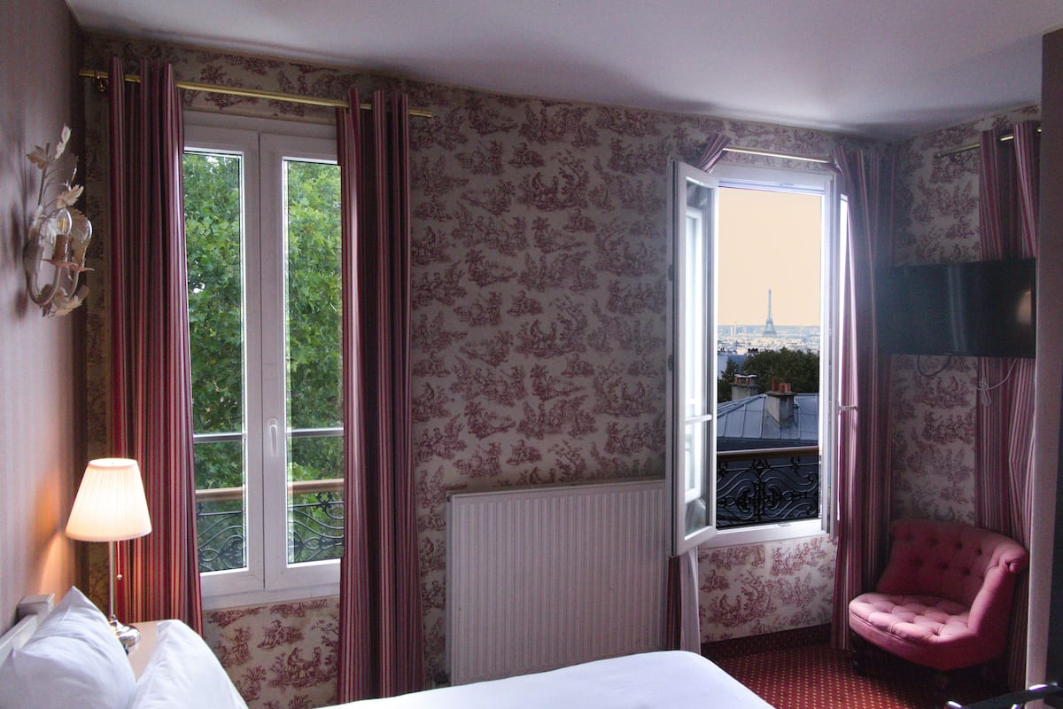 Een hotelkamer met een bed, een stoel, een televisie en een raam met uitzicht op de Eiffeltoren.