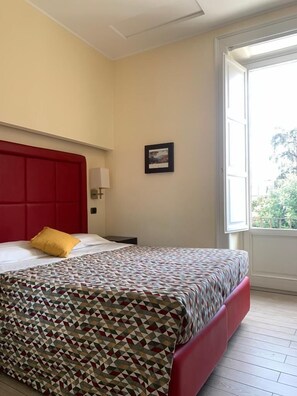 Habitación de hotel con cabecera roja, colcha estampada, almohada amarilla, mesita de noche, lámpara de pared y ventana con vistas a zonas verdes.