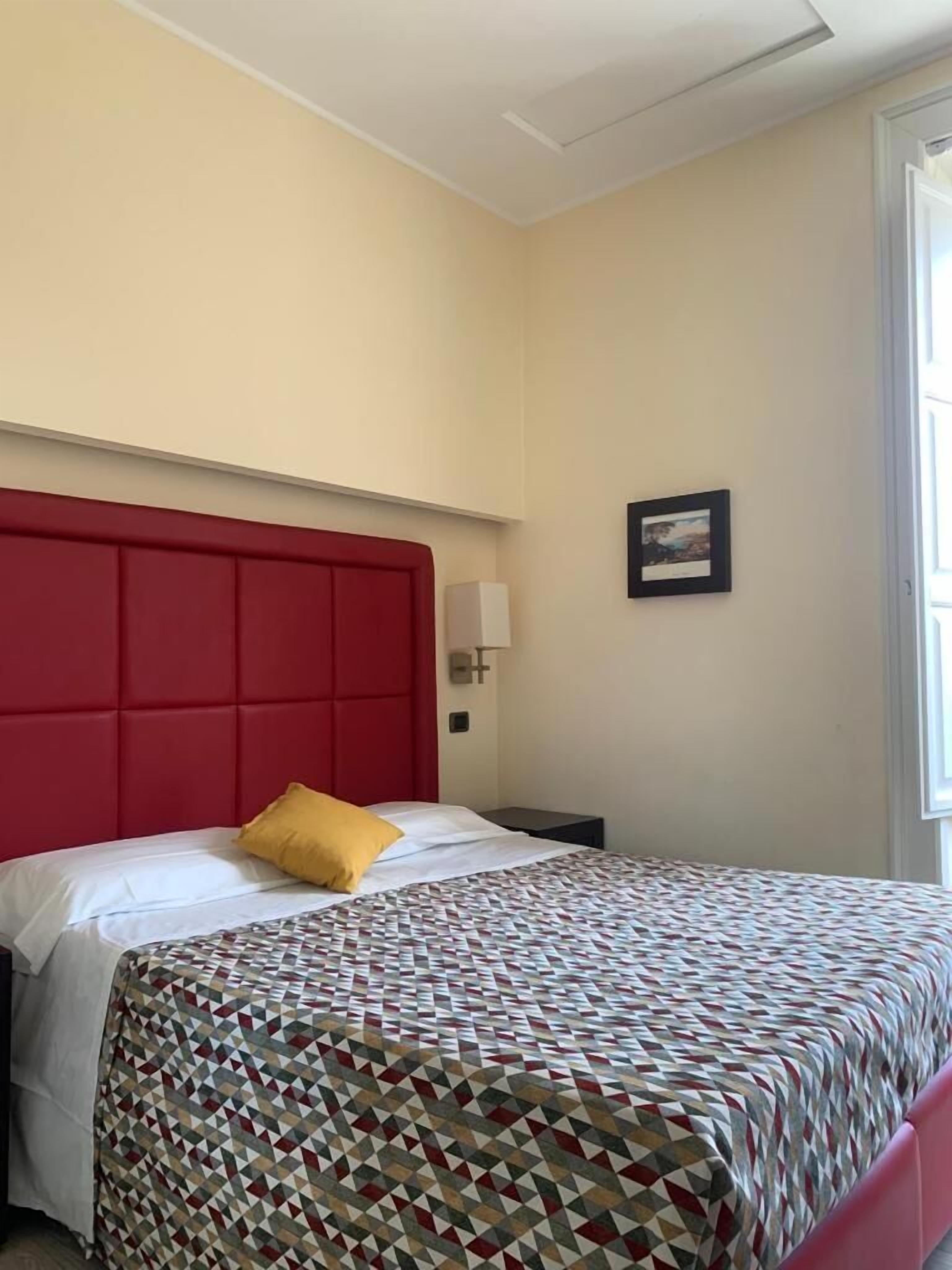 Et hotelværelse med rødt sengegavl, et mønstret sengetæppe, en hvid pude, et natbord med lampe og et billede i ramme på væggen.