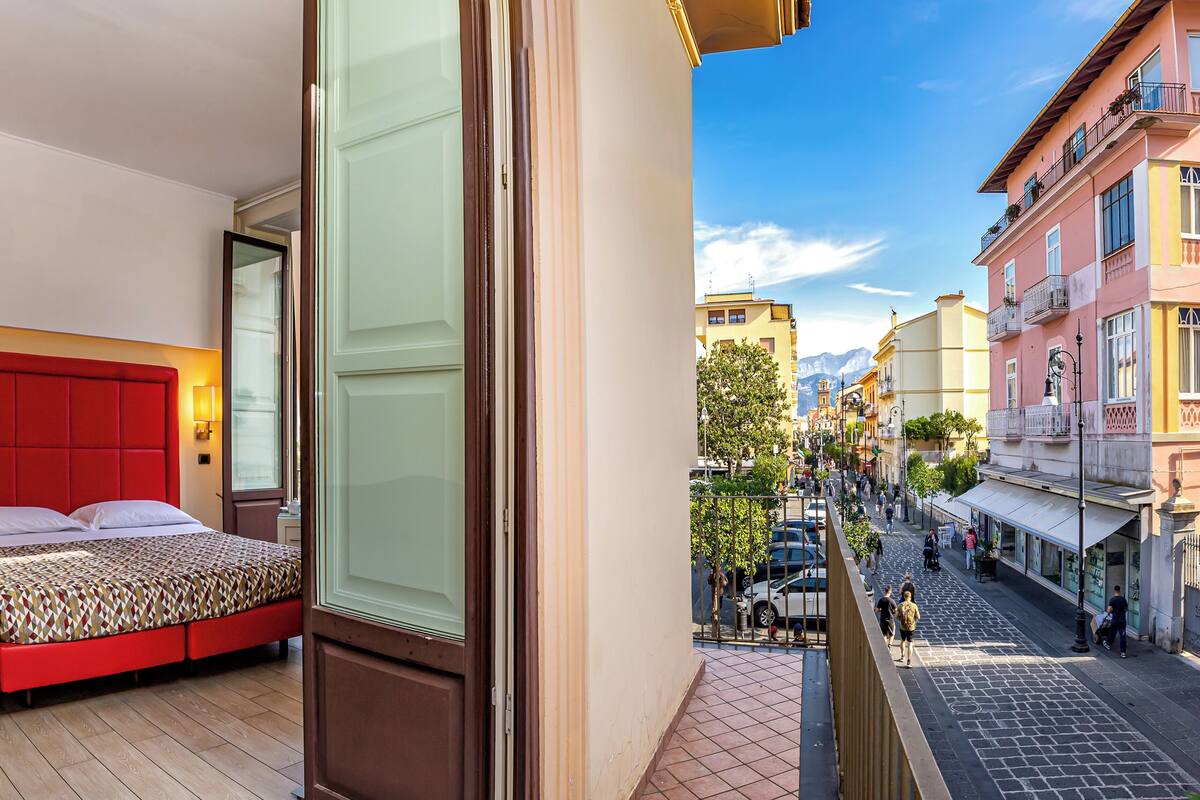 Habitación de hotel con cabecera roja, colcha estampada y vista a una calle concurrida, con peatones y comercios.