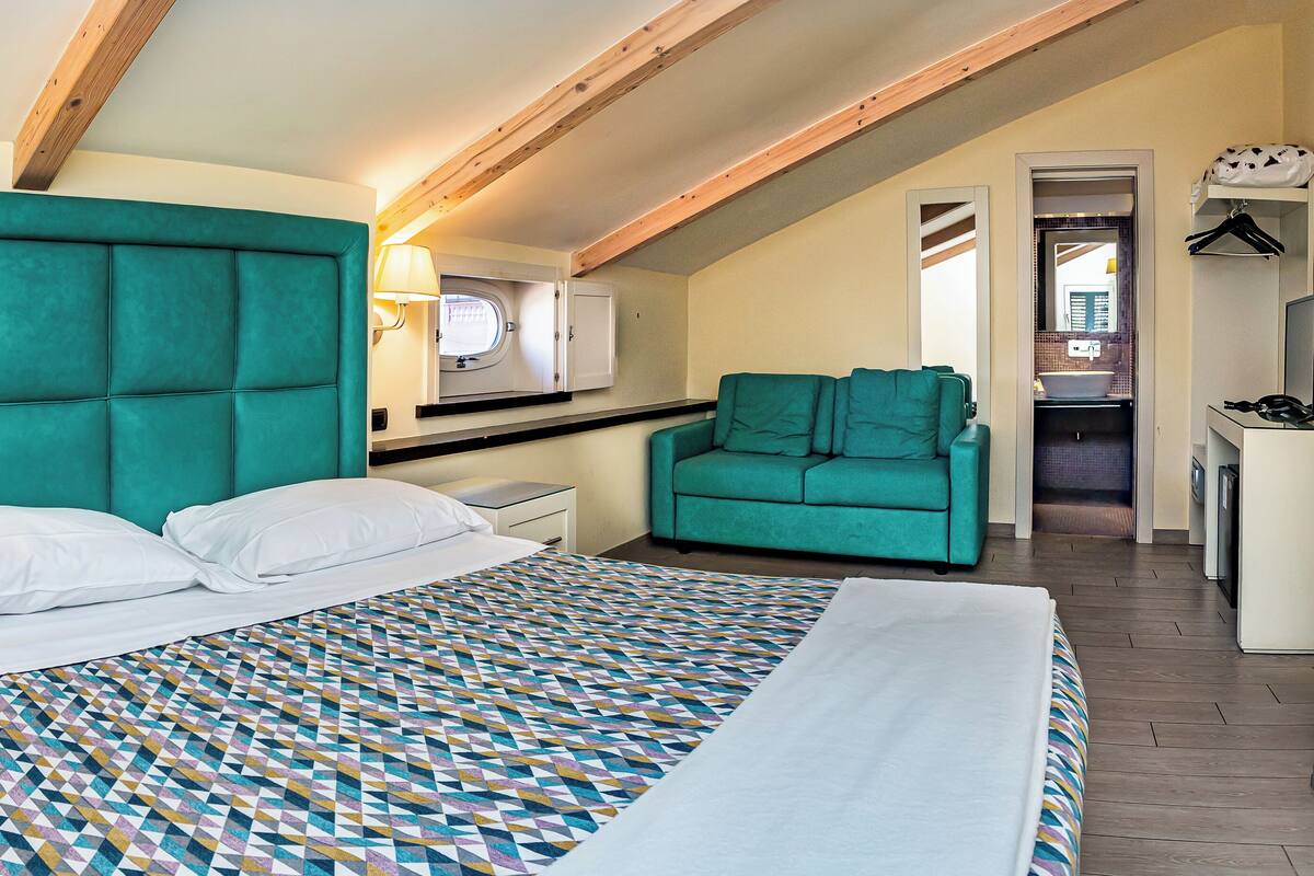 Un dormitorio con una cama grande, cabecera color turquesa, colcha estampada, mesita de noche, cómoda y un sillón color turquesa.