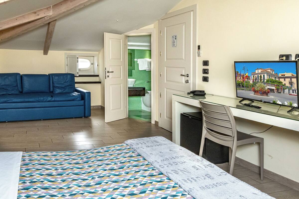 Una habitación compacta con cama, un sofá azul, un escritorio con computadora y un televisor mostrando un paisaje urbano.