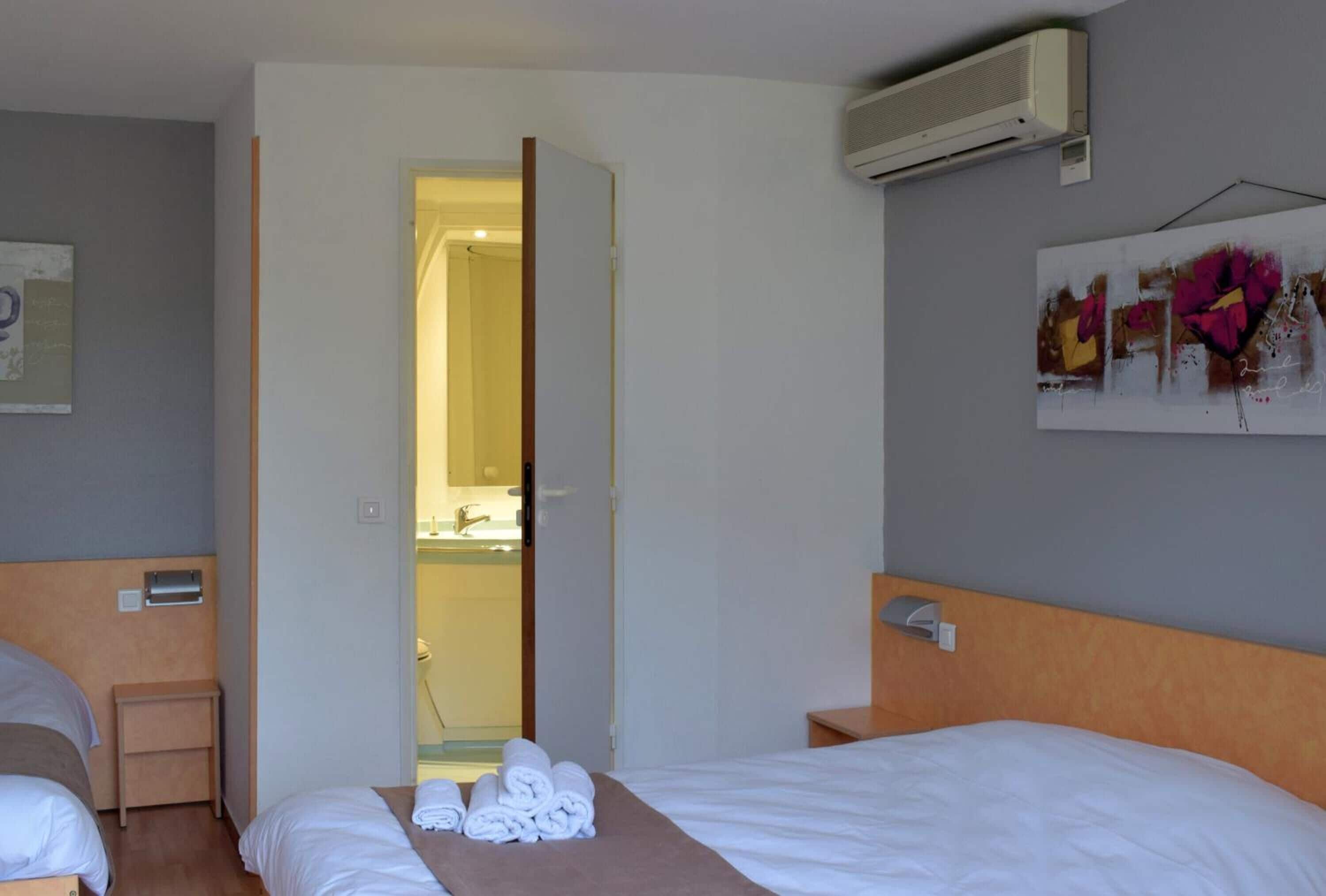 A hotel room with two beds, a bathroom visible through an open door, and a wall-mounted air conditioner.