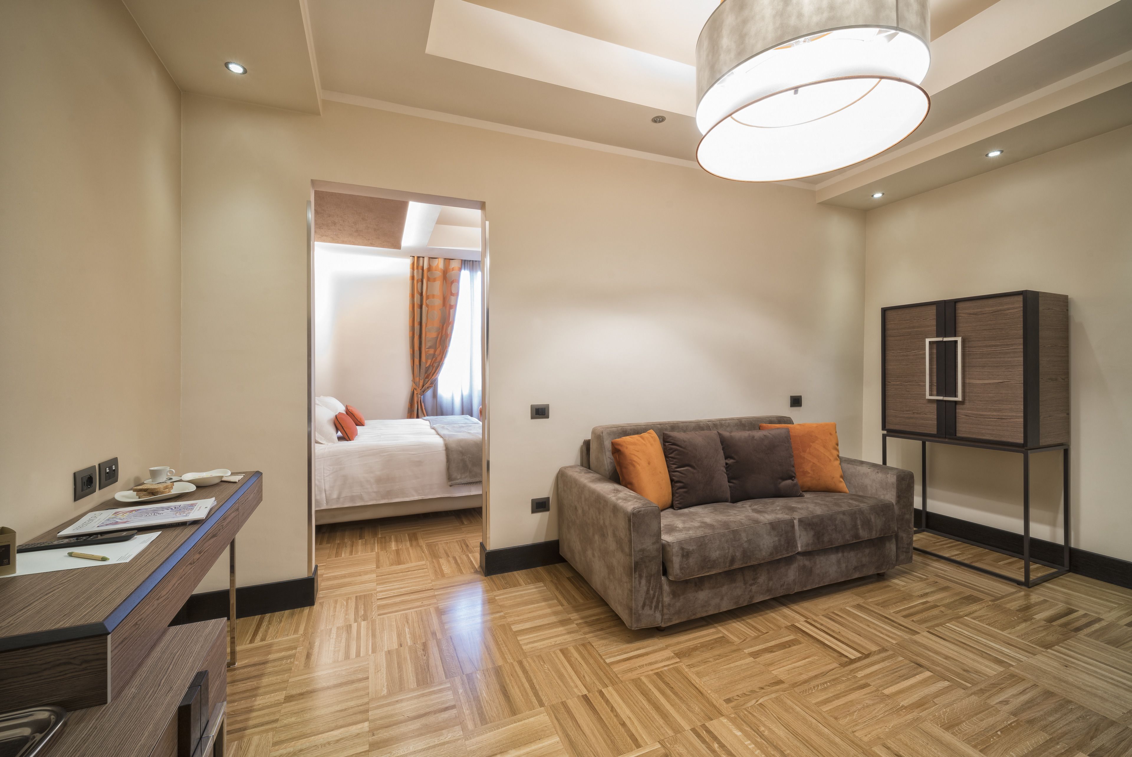 A modern hotel room with a wooden floor, a sofa with orange cushions, a desk with a cup and papers, and a cabinet.
