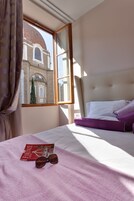 Camera d'albergo con un letto, una finestra che si affaccia su un edificio, un libro e occhiali da sole sul letto.