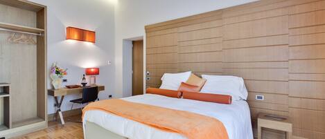 A modern hotel room with a large bed, a desk with a chair, a wardrobe, and a bedside table.