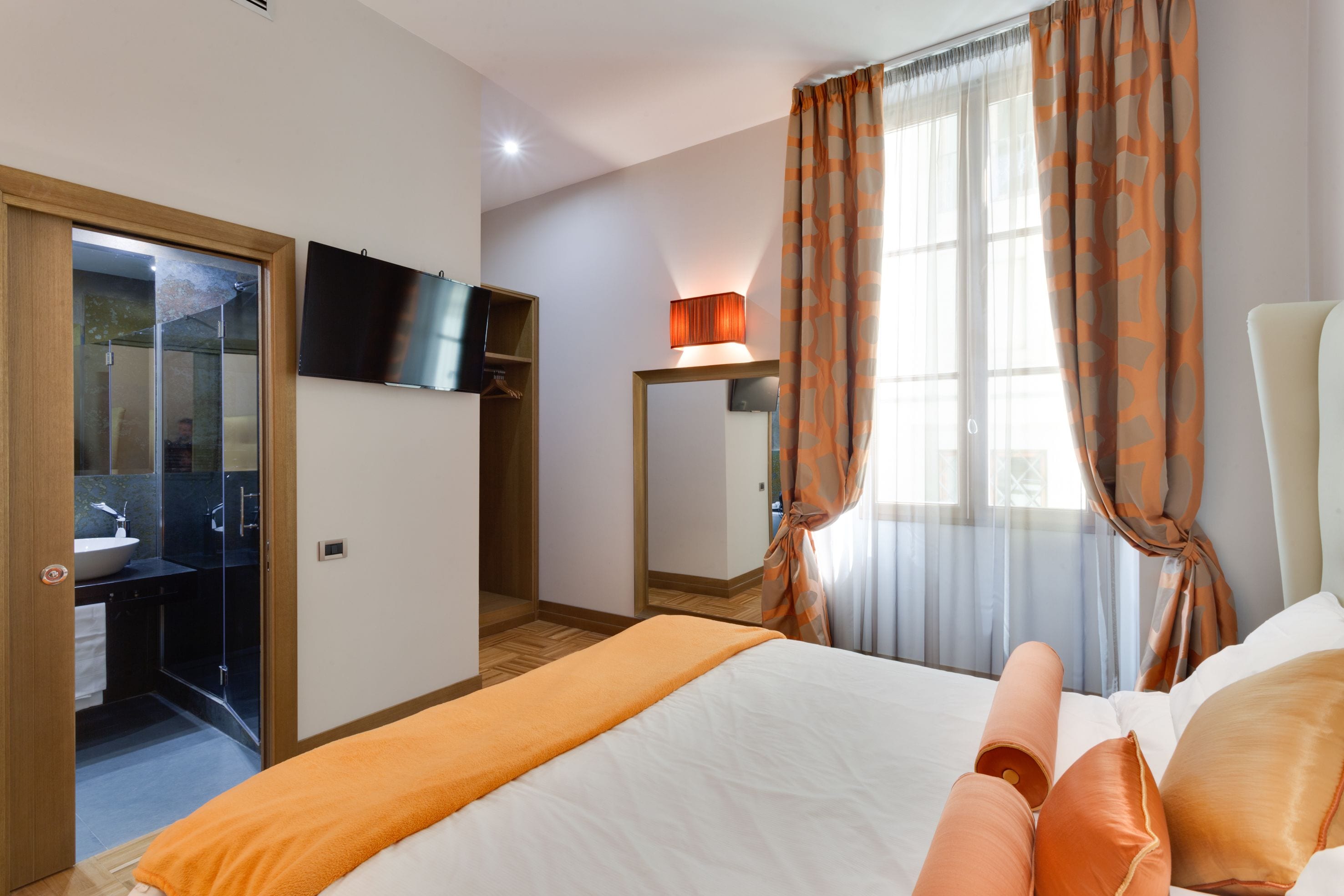 Ein Hotelzimmer mit Bett, Badezimmer mit Dusche, einem an der Wand montierten Fernseher und einem Fenster mit Vorhängen.