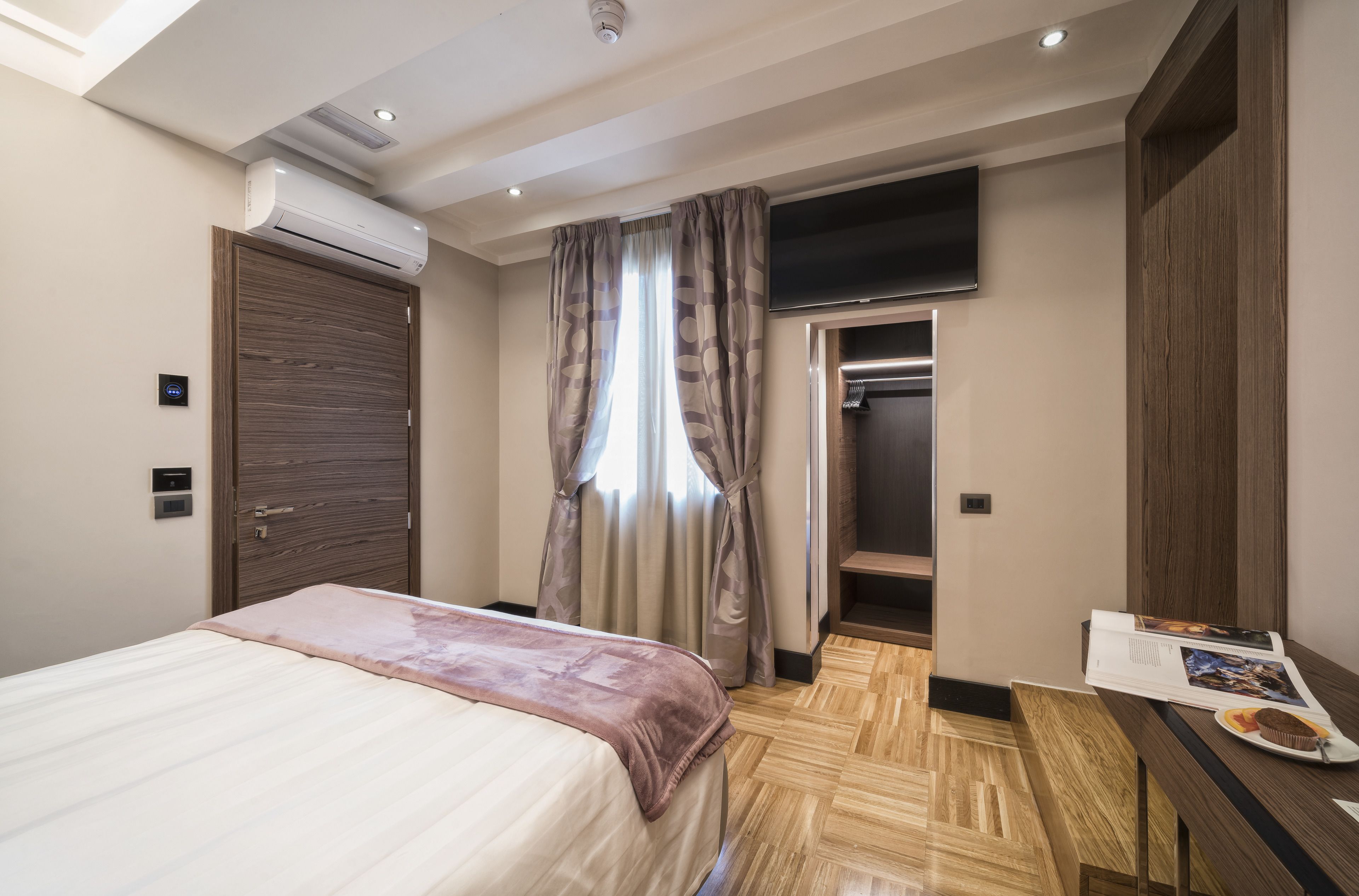 Una habitación de hotel moderna con cama, armario, televisión y una mesita con una revista y un plato.