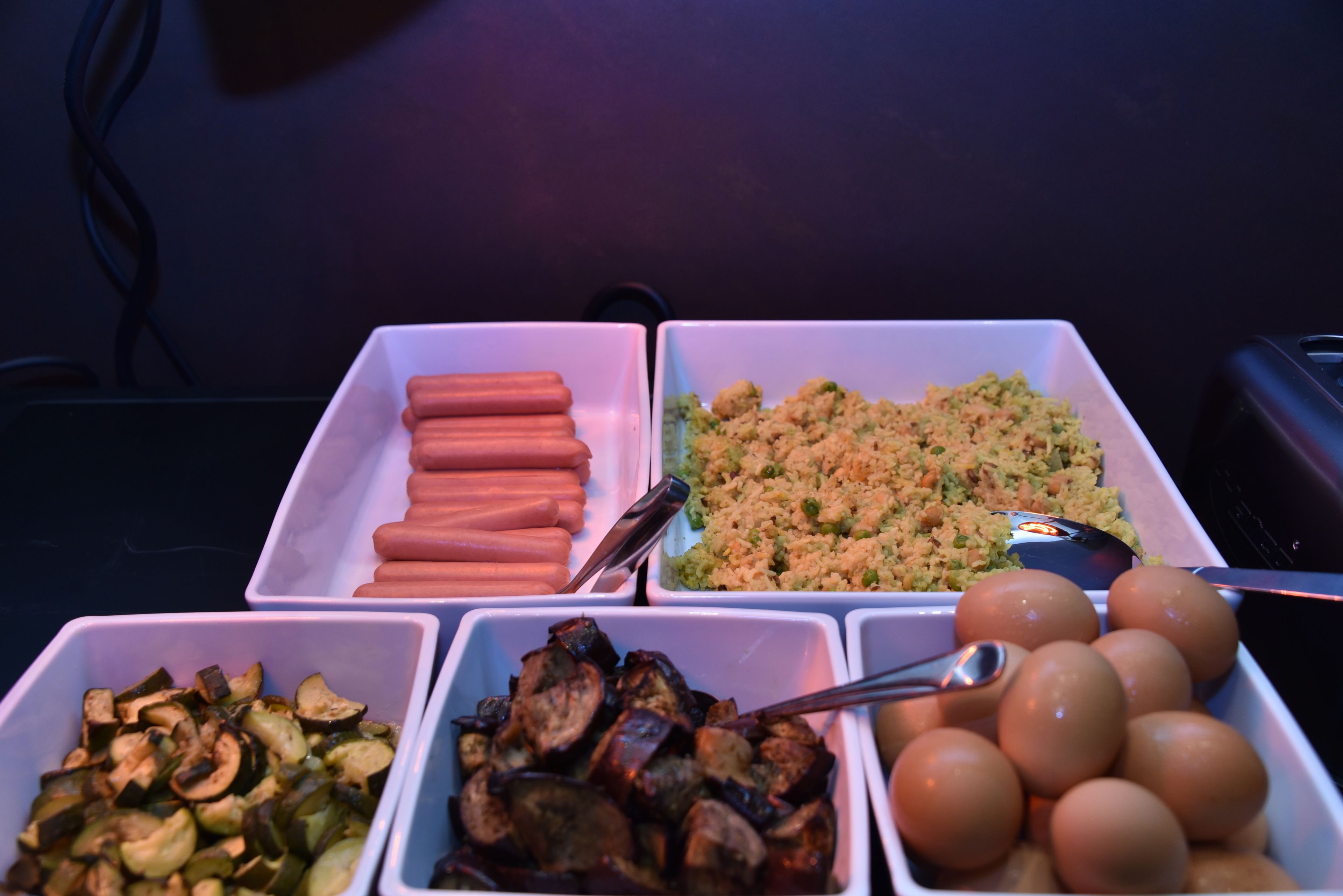 Uma mesa de buffet com diversos alimentos, incluindo cachorros-quentes, arroz, legumes e ovos.