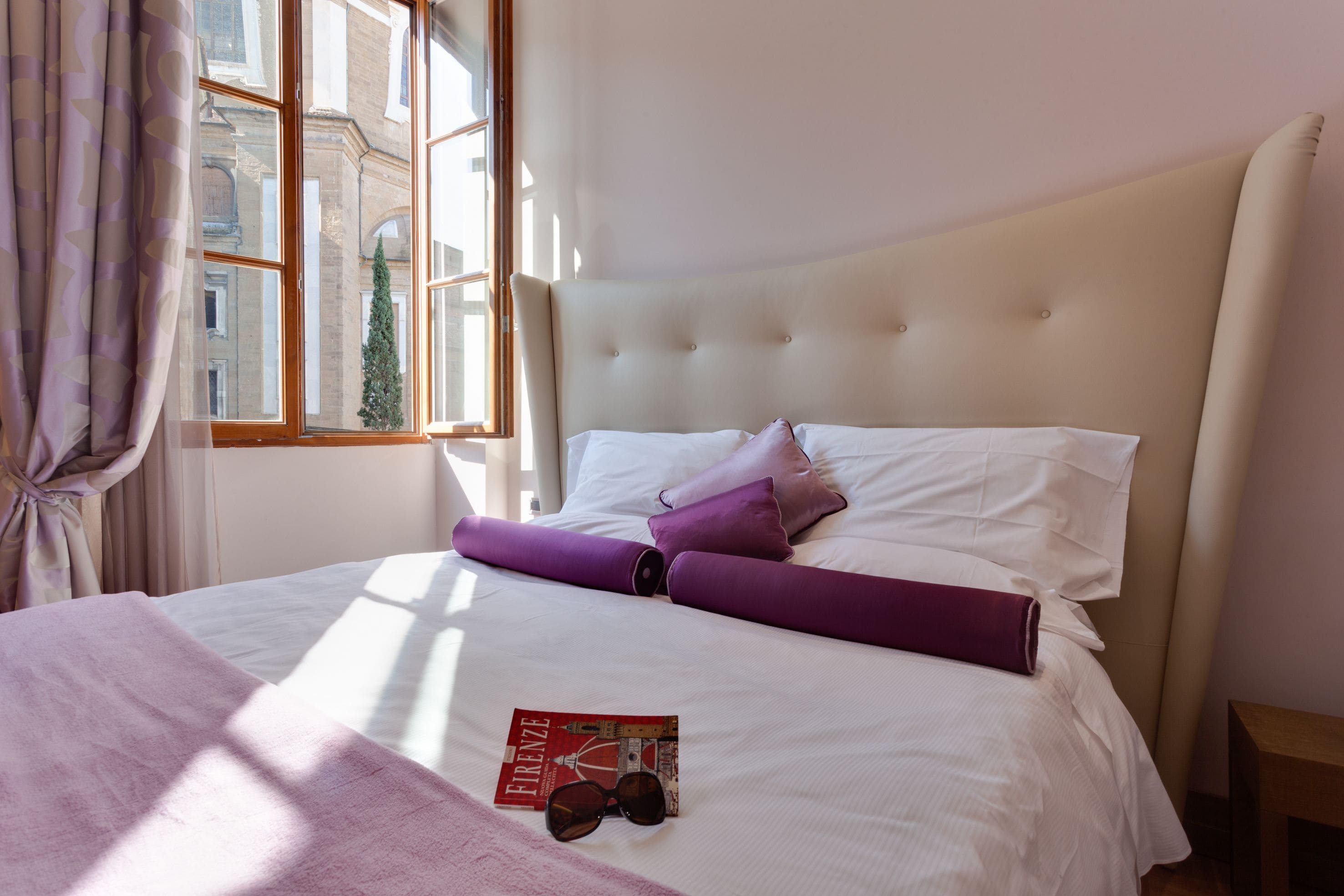 A neatly made bed with white and purple bedding, a magazine, and sunglasses on the bed.