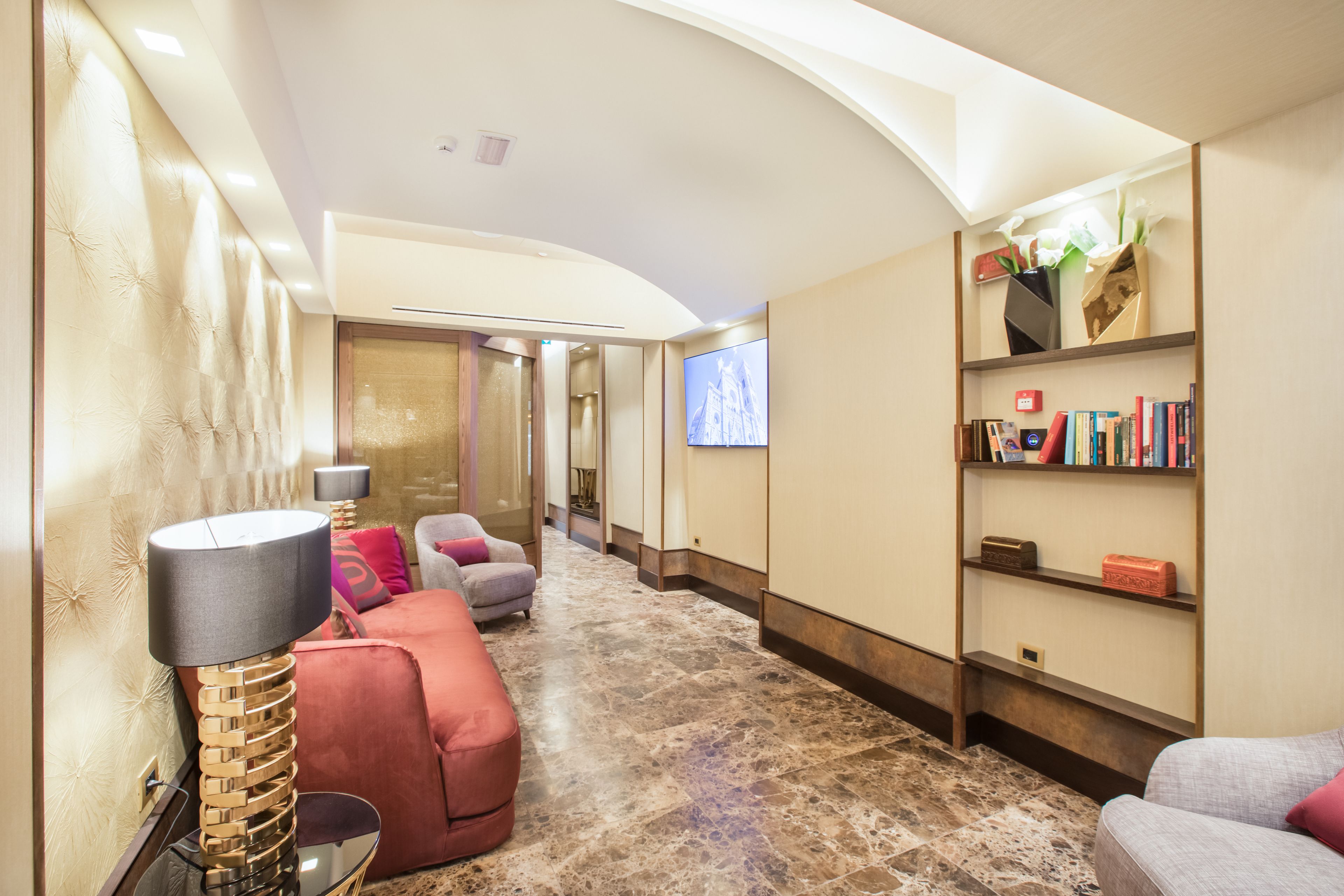A spacious hallway with a plush red sofa, a gold lamp, and a bookshelf with books and decorative items.