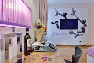 Una stanza con una scrivania, una macchina da scrivere, una bottiglia di vino, un bicchiere di vino, un bouquet di fiori, una sedia e un muro con una televisione a muro e elementi decorativi.