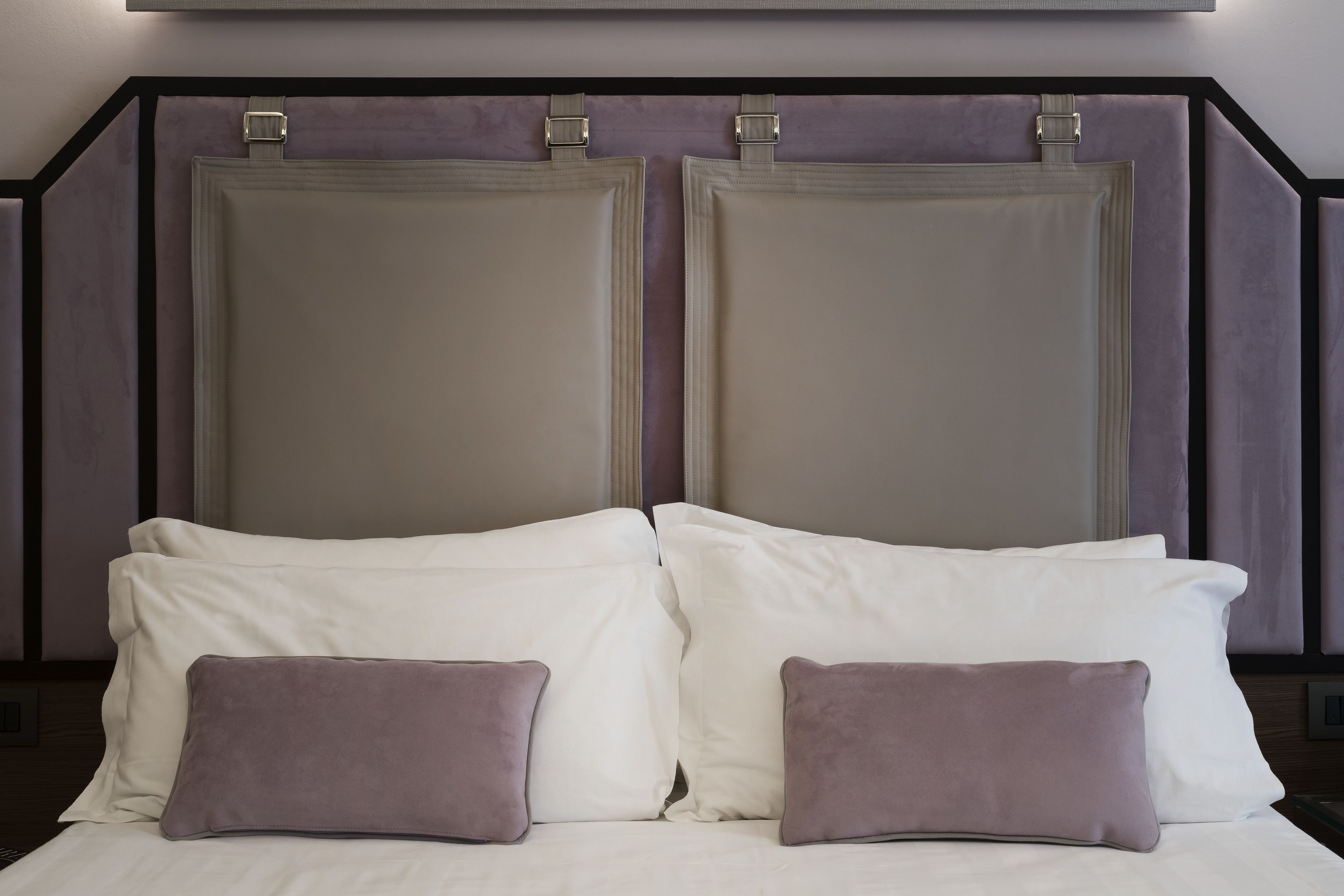 Un letto con lenzuola bianche e due cuscini viola, una testiera con struttura metallica e un muro con l'interruttore della luce.