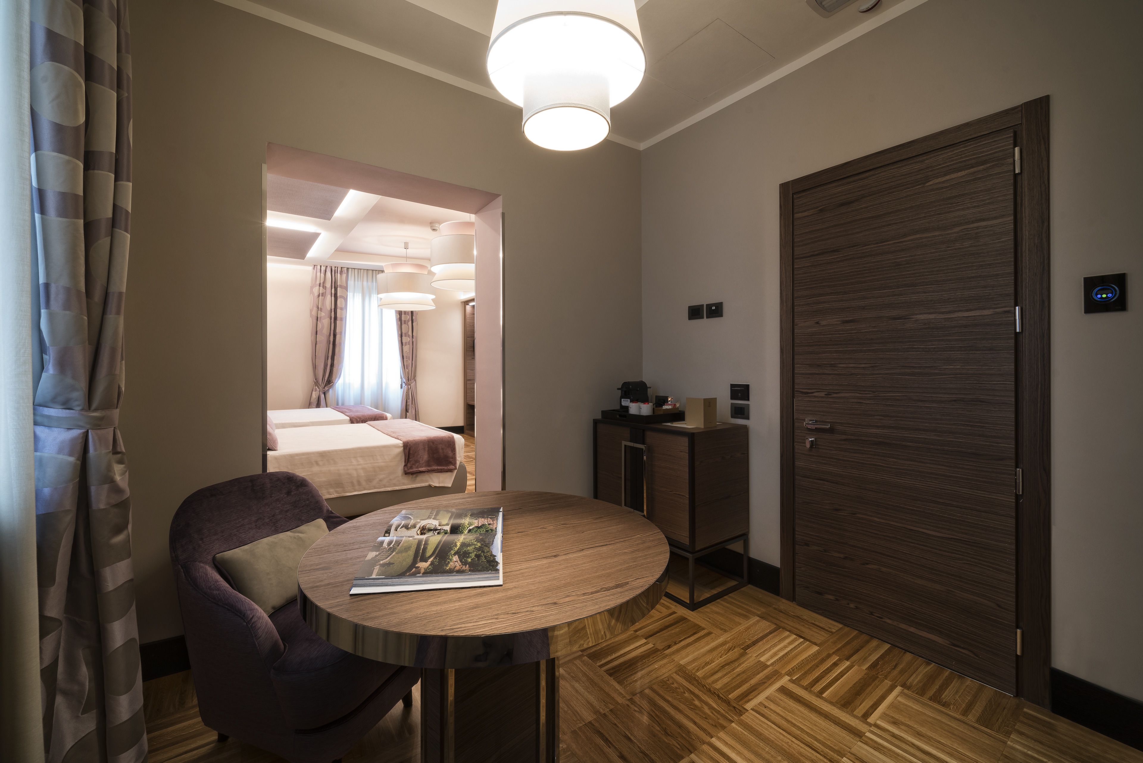 A hotel room with a wooden floor, a round table with a magazine, a purple chair, a large wooden door, and a view into another room with a bed and curtains.