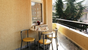 A balcony with a table and chairs, a window with blinds, and a view of trees and buildings. - Hotel Victoria (Skopje)