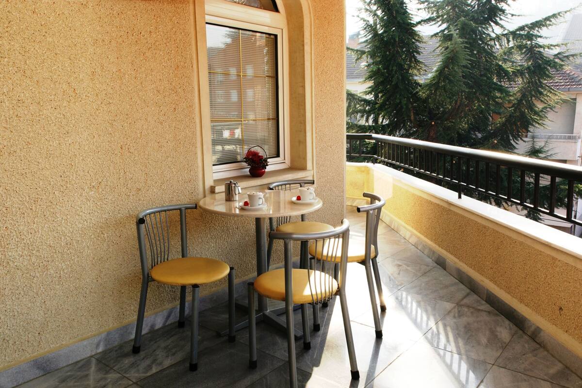 Un balcón con mesa y sillas, una ventana con persianas, y vistas a árboles y edificios.