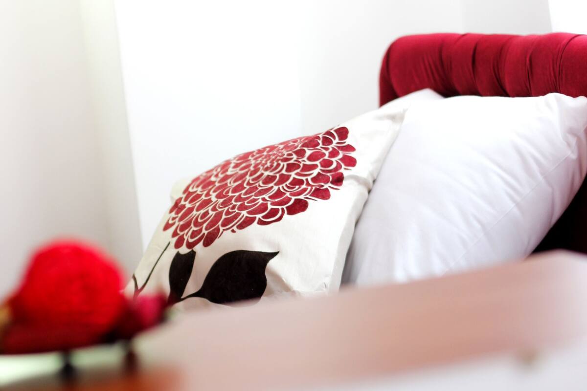 Una cama con cabecera tapizada en rojo, ropa de cama blanca y un cojín decorativo con estampado floral rojo.