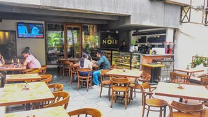 An outdoor cafe with wooden tables and chairs, a TV mounted on the wall, and a sign for 'NOIR'. - Kings International Juhu (Mumbai)