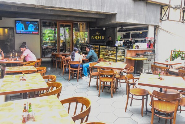 An outdoor cafe with wooden tables and chairs, a TV mounted on the wall, and a sign for 'NOIR'. - Kings International Juhu (Mumbai)