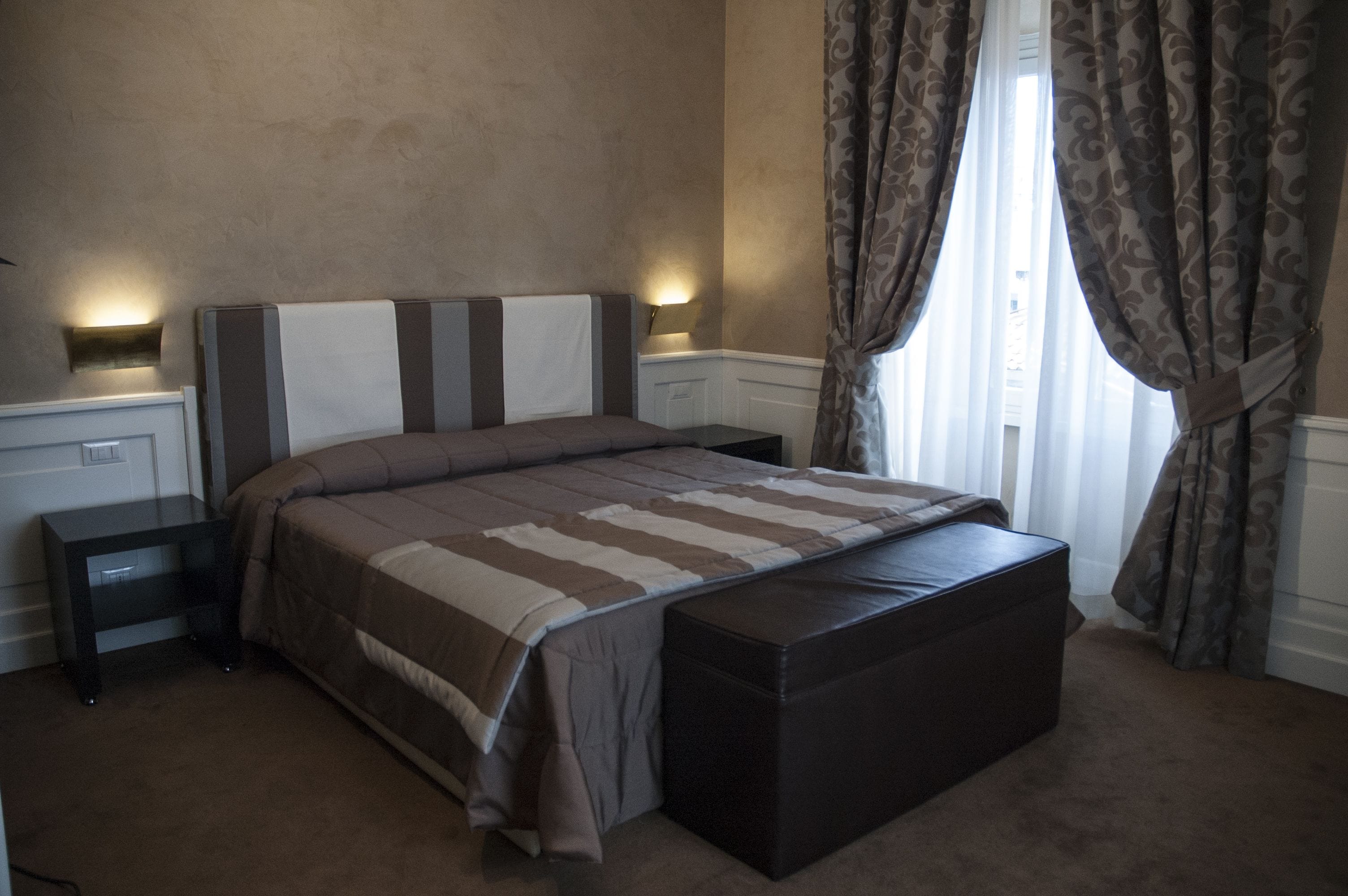 A bedroom with a large bed, a nightstand, a bench, and a wall-mounted light.