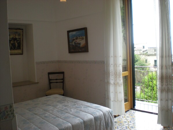 A bedroom with a bed, a chair, and a balcony with a view of buildings and greenery. - L'Argine Fiorito (Atrani)