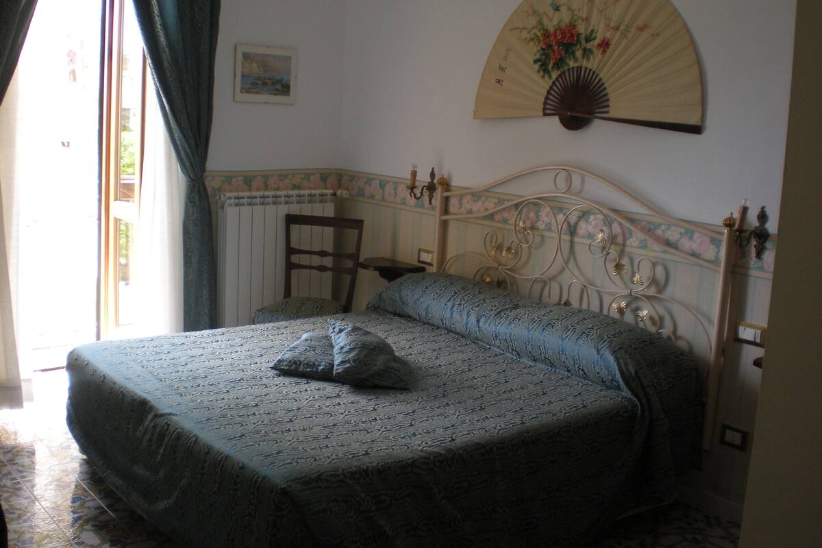 A bedroom with a bed, a chair, a fan, and a window with curtains.