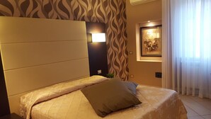 A hotel room with a bed, a headboard, a wall-mounted light, a framed picture, and a window with curtains. - B&B Emozioni Romane (Rome)