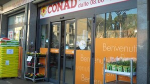 A storefront with the sign 'CONAD' and a flower logo. There are orange welcome signs on the windows, and a stack of yellow bags outside. - B&B Emozioni Romane (Rome)