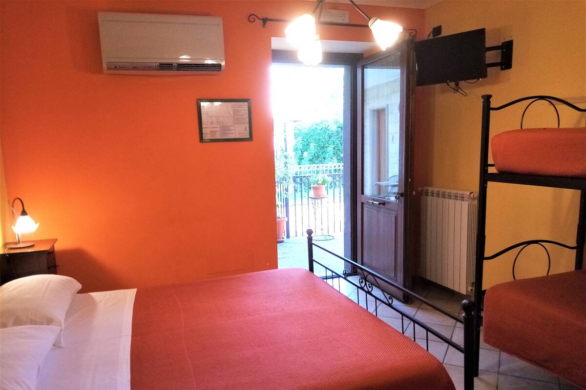 A room with a bed, a bedside table with a lamp, a bunk bed, a window with a view of the outside, and an air conditioning unit on the wall.