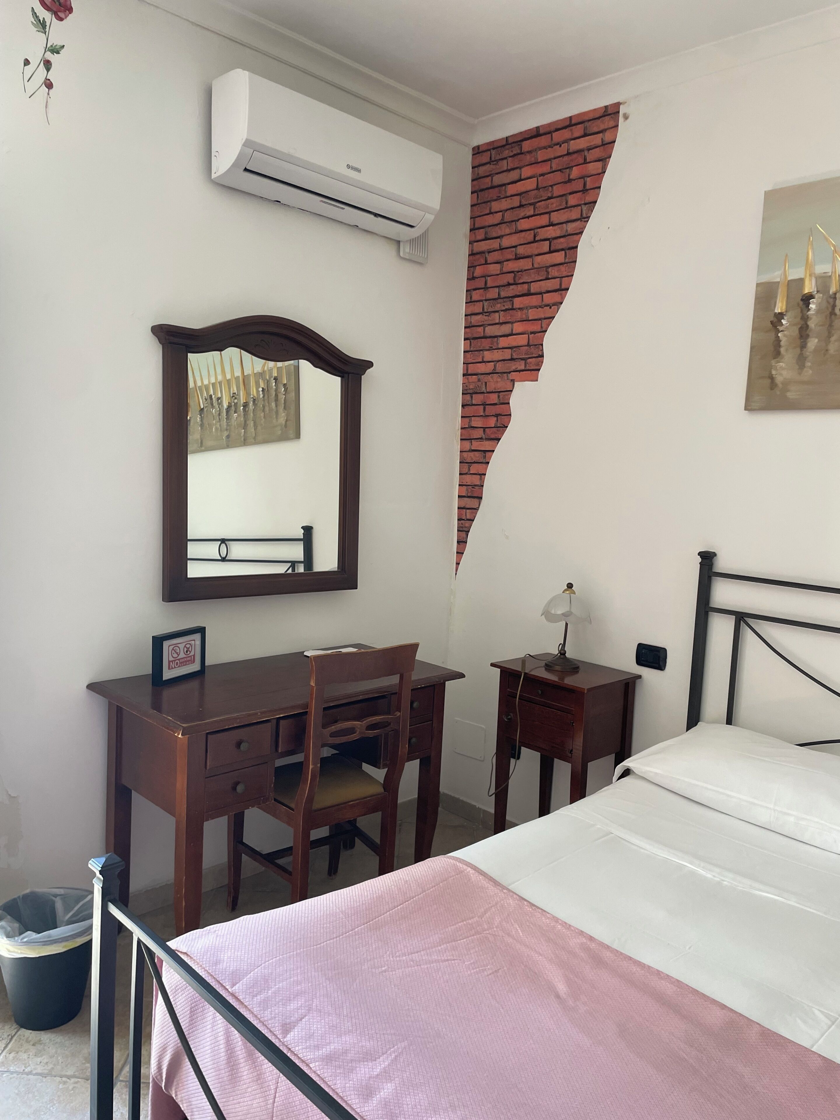 A bedroom with a bed, a desk with a chair, a small table with a lamp, a wall-mounted air conditioner, and a partial brick wall.