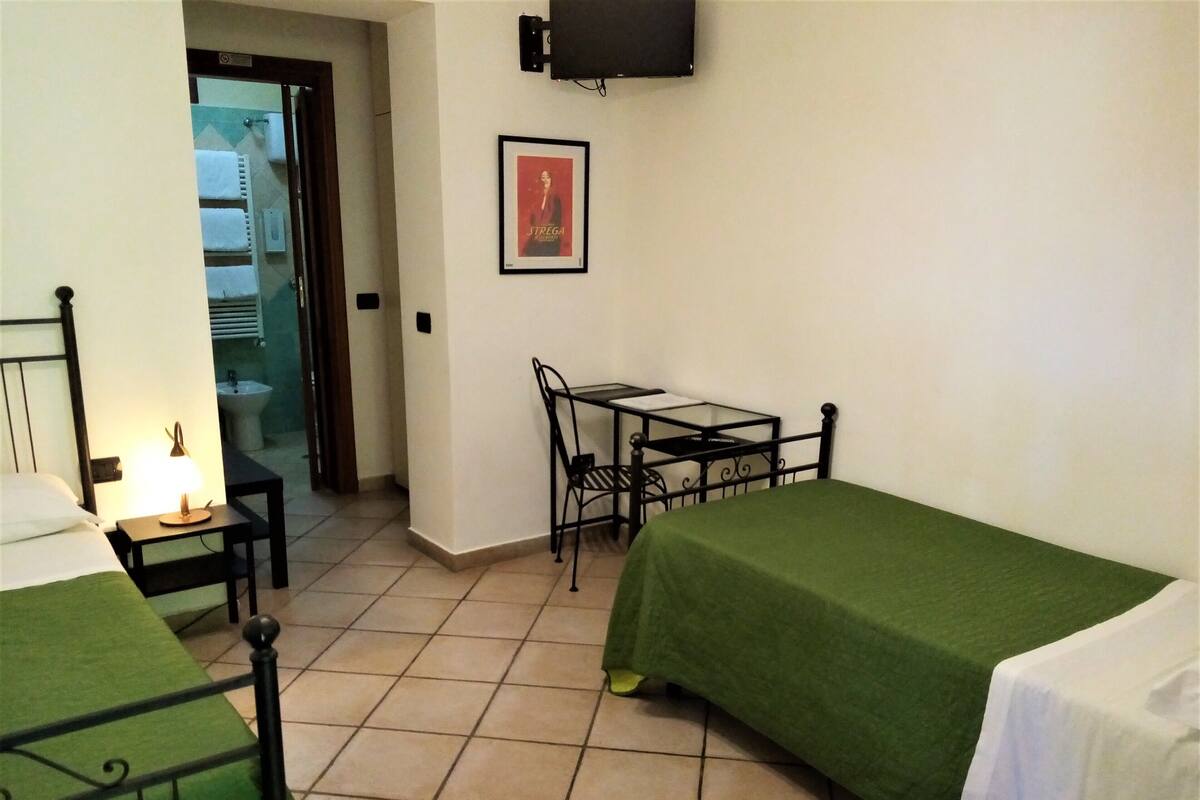 A hotel room with two beds, a small table, a chair, a TV, and a bathroom visible through an open door.