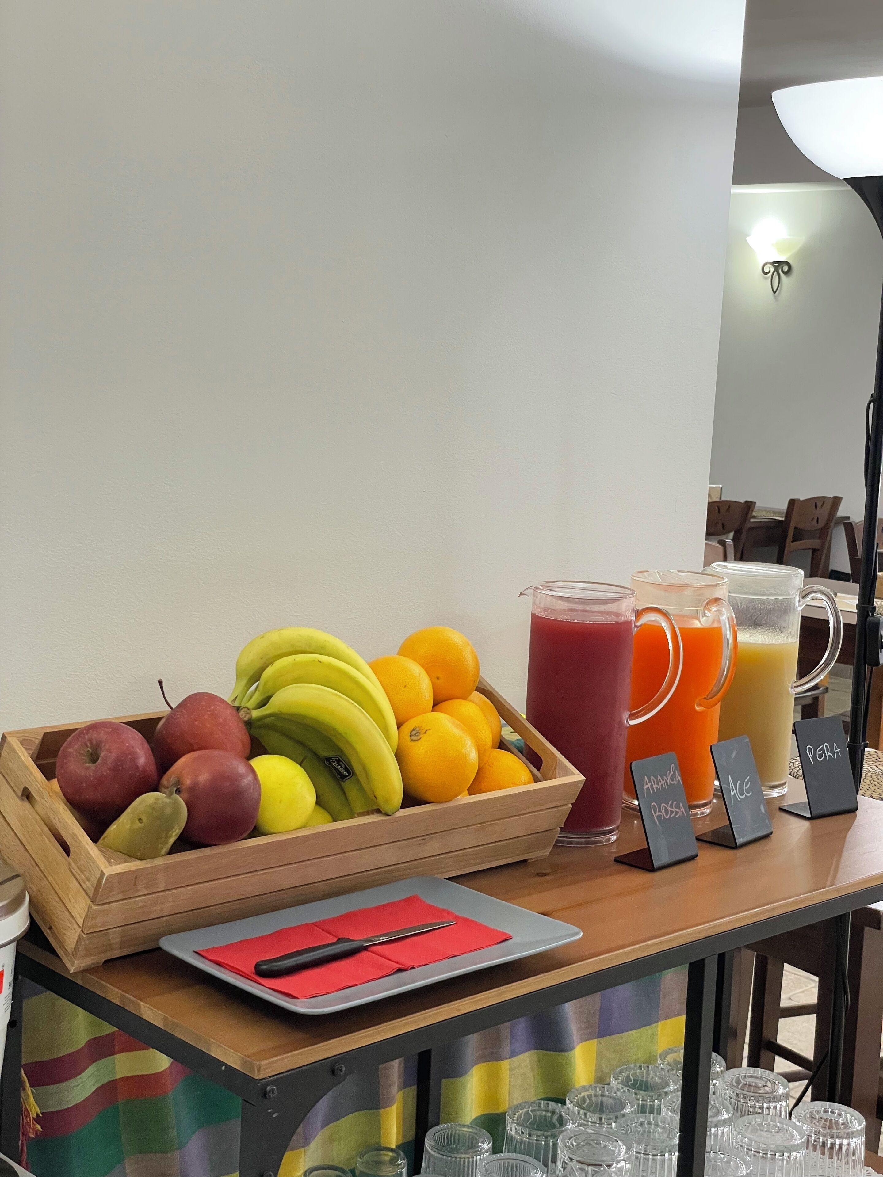 Una caja de madera con manzanas, plátanos y naranjas, una bandeja con servilleta roja y cuchillo, y tres jarras etiquetadas con jugos.