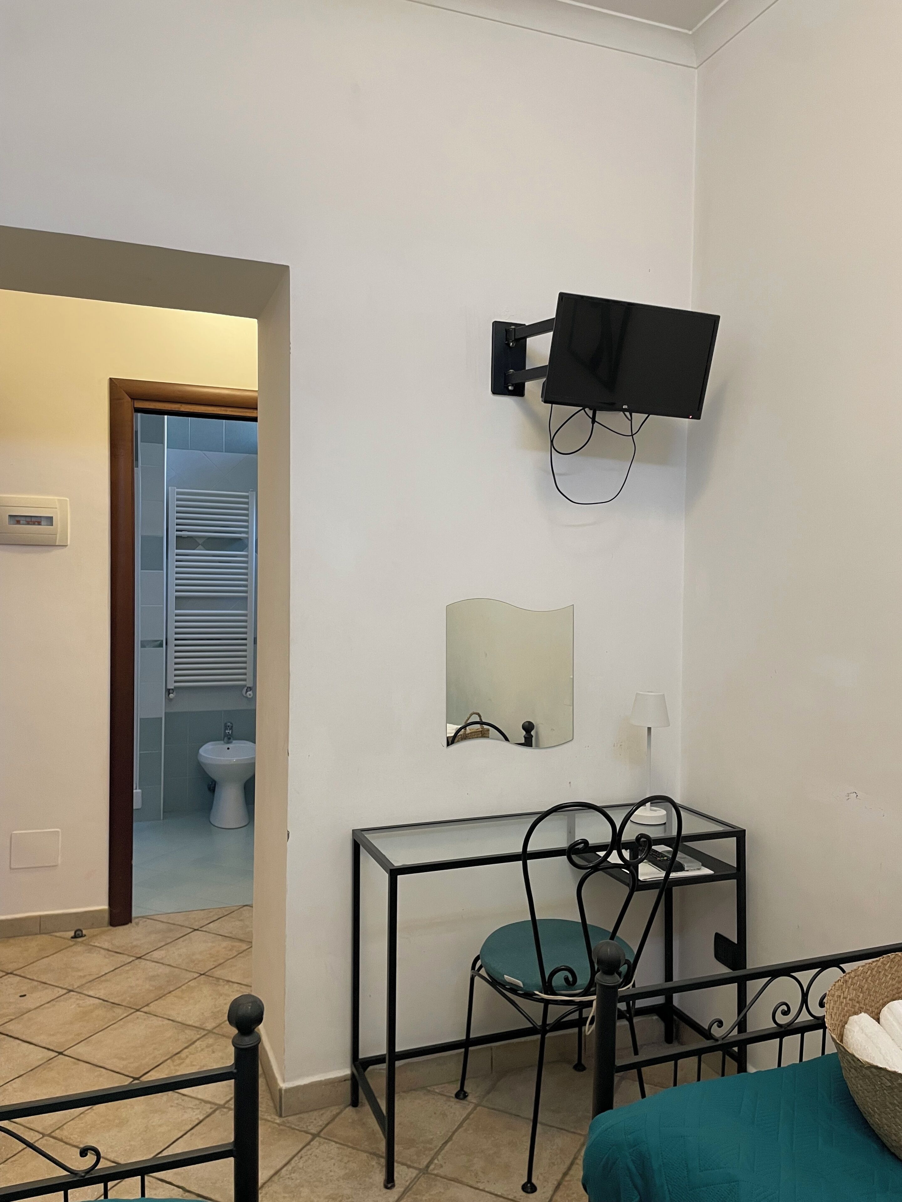 A room with a bed, a desk with a chair, a television mounted on the wall, a mirror, and a bathroom visible through an open door.