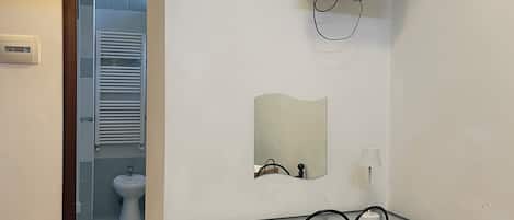 A room with a bed, a desk with a chair, a television mounted on the wall, a mirror, and a bathroom visible through an open door.