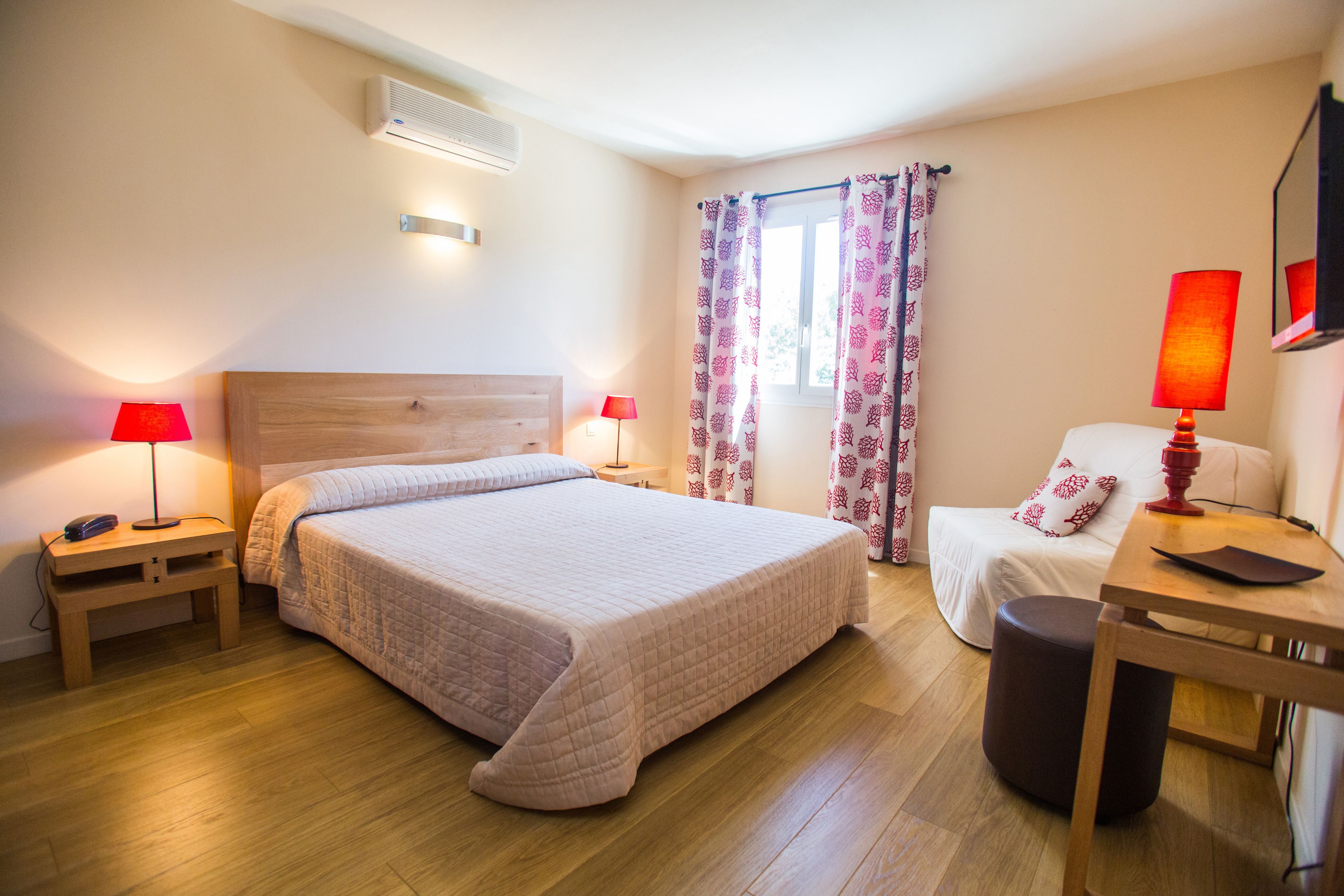 Une chambre d’hôtel avec un grand lit, des tables de chevet avec des lampes, un bureau en bois et une fenêtre agrémentée de rideaux à motifs.