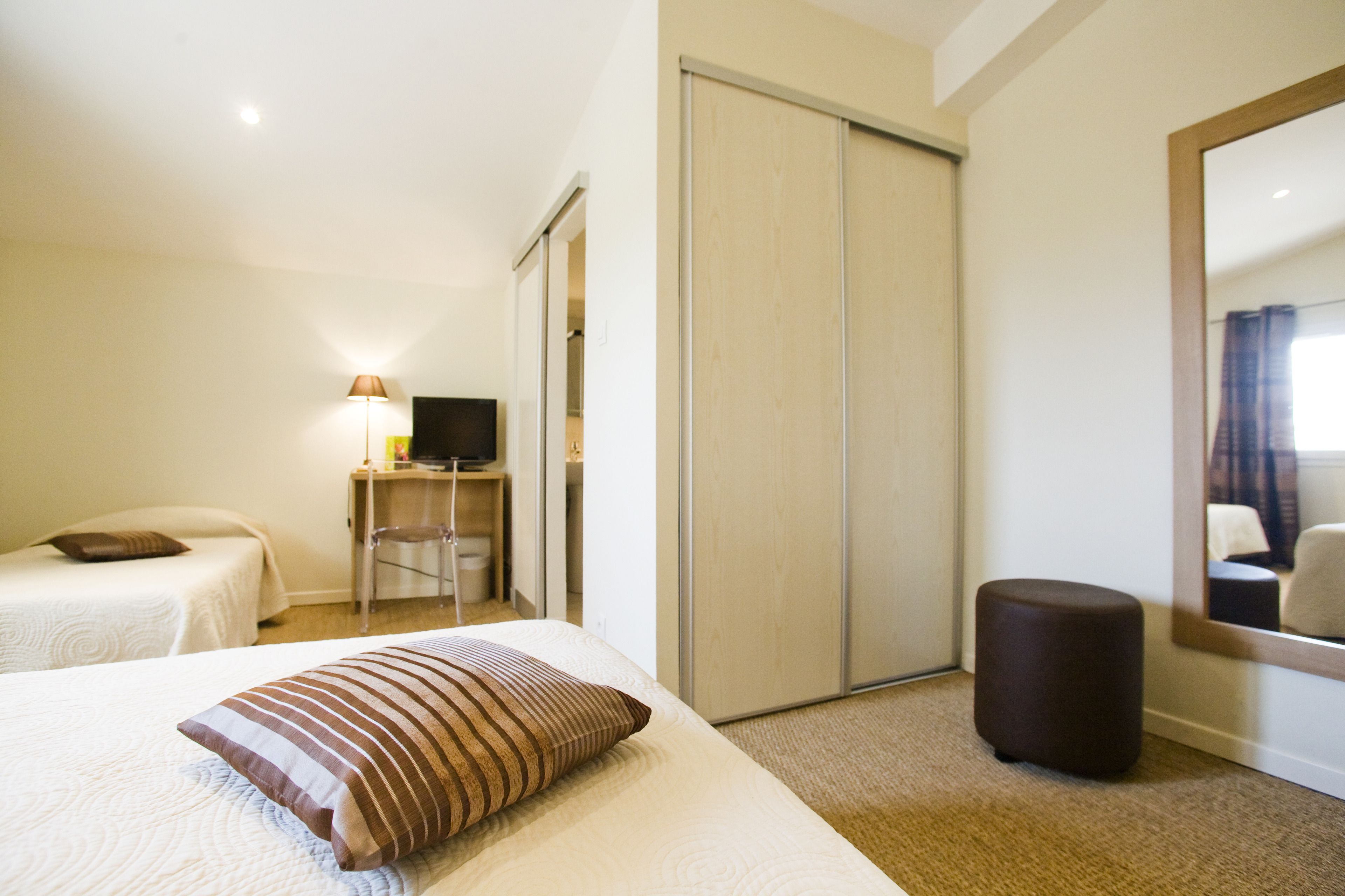 A hotel room with two beds, a desk with a chair, a TV, a wardrobe, and a small ottoman.
