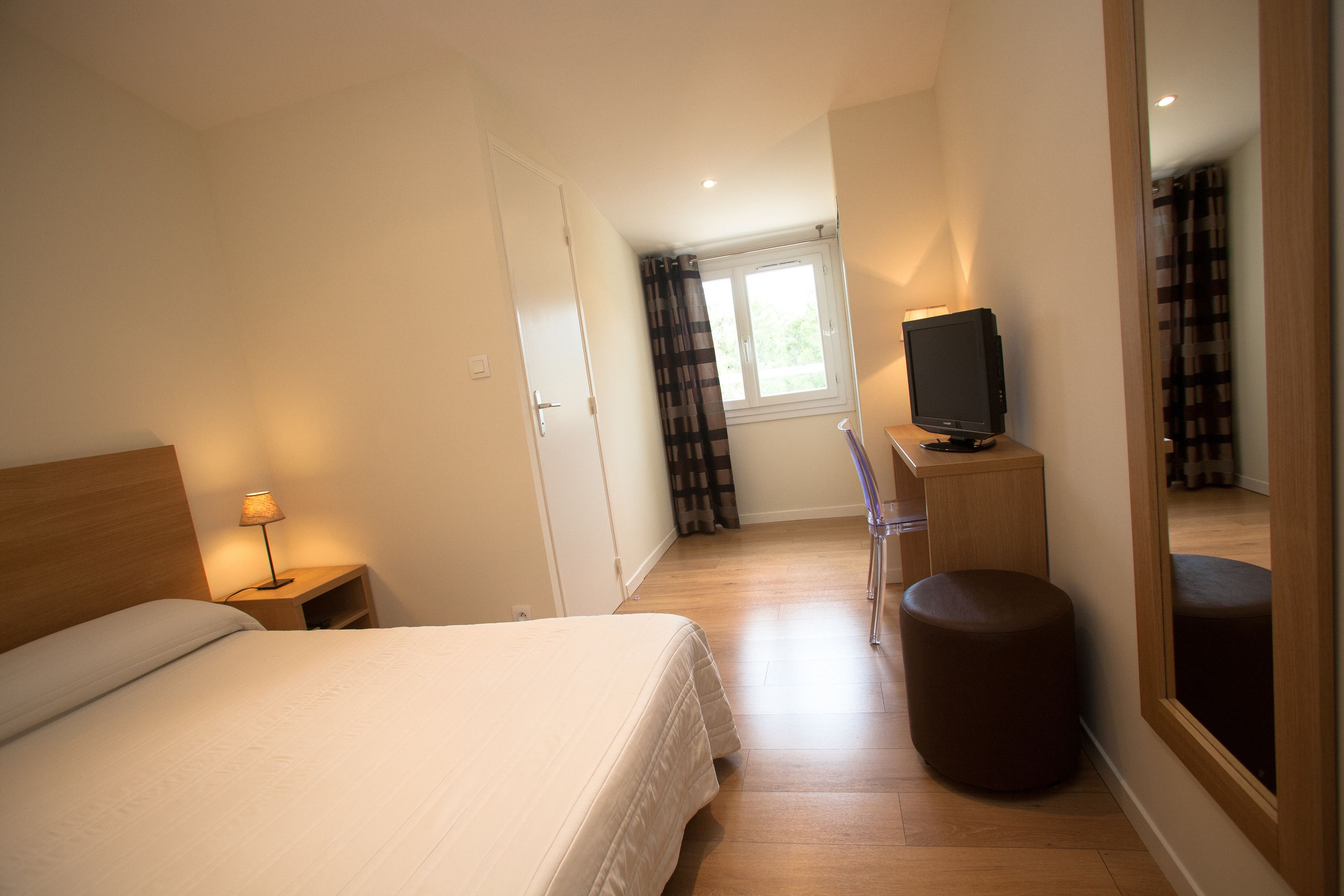 A hotel room with a bed, a desk with a computer, a chair, a small stool, and a television.