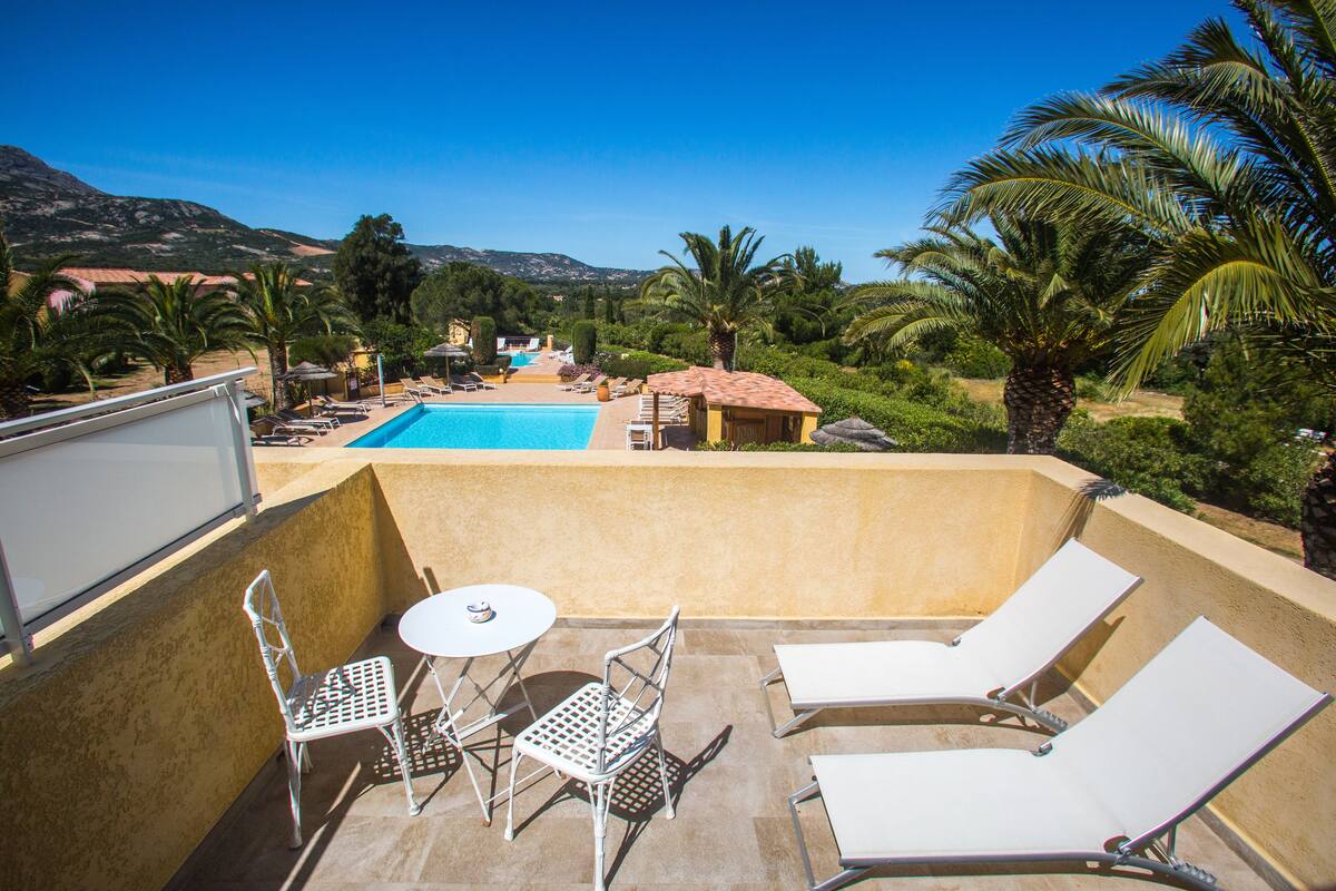 A rooftop pool area with lounge chairs, a table, and chairs, overlooking a pool and a mountainous landscape.