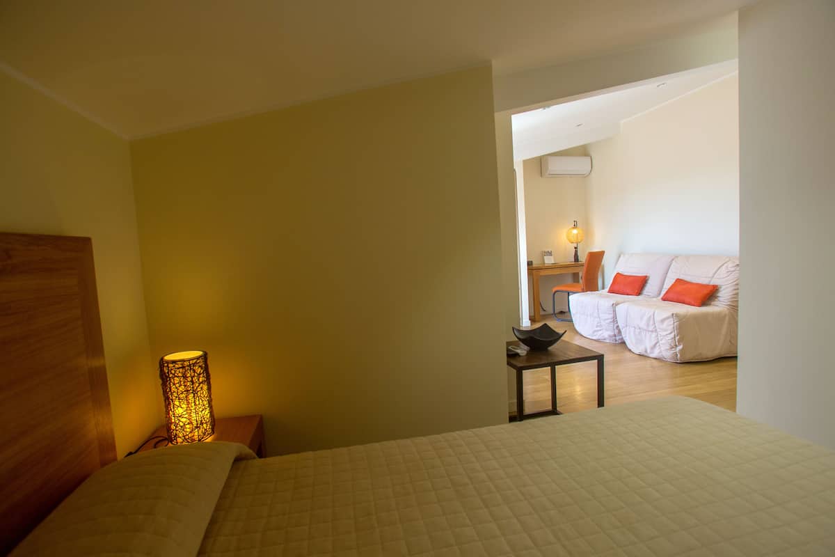 A bedroom with a bed, bedside table, lamp, and a view into another room with a sofa and a table.